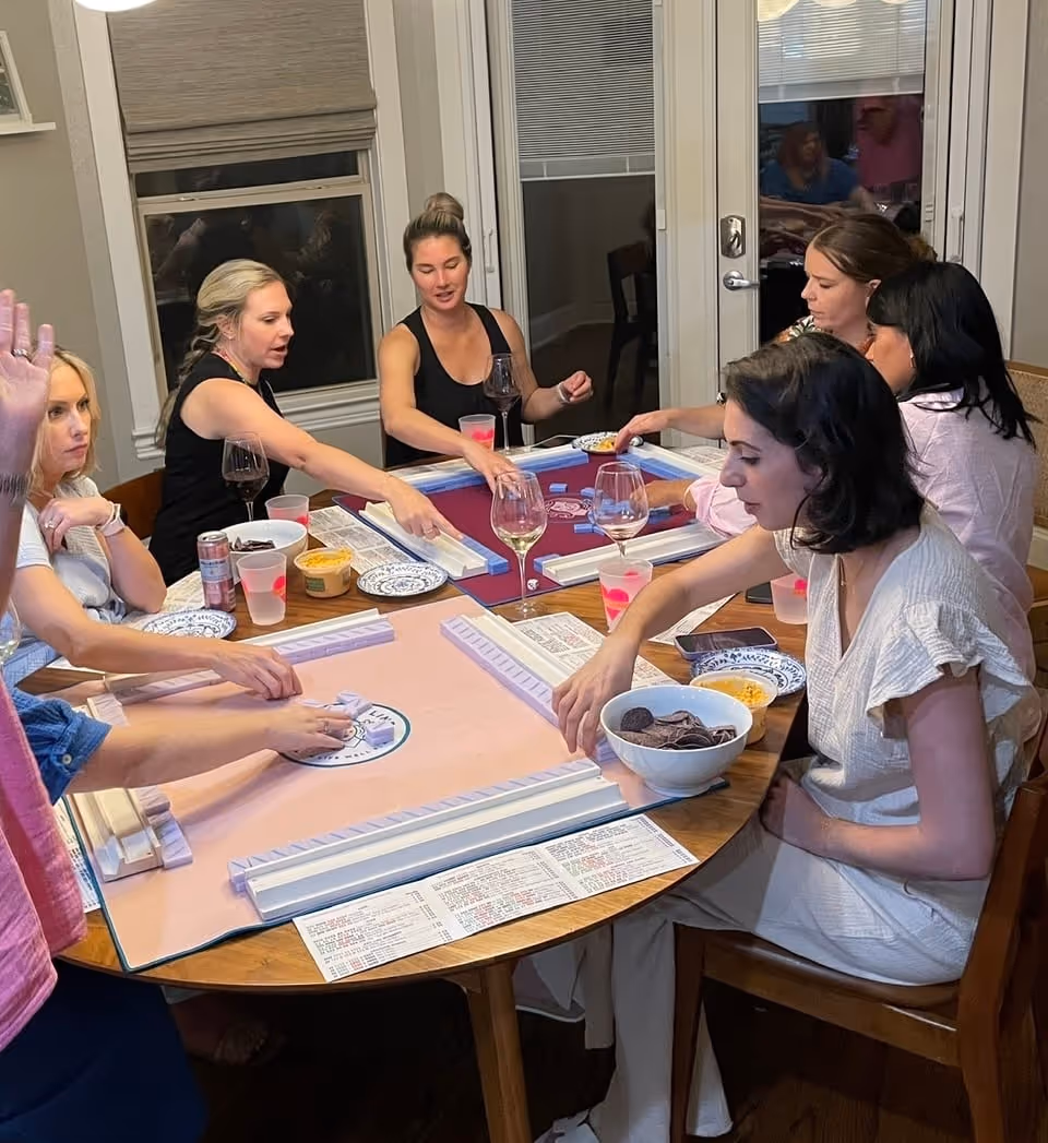 Friends learning American mahjong around a dining table at home, tiles and racks in front of them